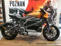Pomarańczowy harley elektryczny z czarnymi akcentami, prezentowany na drewnianej podłodze, z plakatem Harley-Davidson w tle.