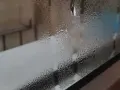 Zaparowane okna od zewnątrz, z kroplami wody tworzącymi wzory. W tle widać niewyraźne kształty.