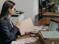 Kobieta w okularach przegląda dokumenty, szukając tytułu prawnego do lokalu. Na biurku leżą też laptop, kalkulator i sterty papierów.