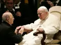 Papież Jan Paweł II w białej szacie, klęczący przed nim mężczyzna w czerni. W tle widać innych duchownych i dziennikarzy, być może z zakonu **jezuitów**.