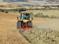 Trattore giallo in azione per l'aratura del terreno, sollevando terra e polvere in un paesaggio rurale collinare.