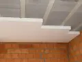 Ocieplenie stropu styropianem w trakcie budowy. Widoczne płyty styropianowe pod sufitem, betonowe belki stropowe i ściany z cegły.