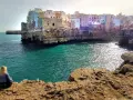 Kobieta siedzi na skale, podziwiając widok na Polignano a Mare. Białe domy wznoszą się na klifie nad turkusowym morzem.
