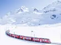 A bright red St. Moritz train winds through a pristine, snow-covered mountain landscape under a clear blue sky.