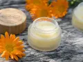 Ringelblumensalbe gegen Falten: Natürliche Pflege für glatte Haut. Kleine Gläser mit Salbe und leuchtend orangefarbene Ringelblumenblüten auf Holz.