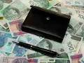 Czarny portfel i długopis leżą na stosie polskich banknotów.