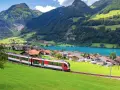 A red and white train travels through a picturesque Swiss village nestled beside a turquoise lake, with lush green mountains in the background. This is the scenic route from Lucerne to Lugano.