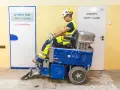 Pracownik w kasku i kamizelce obsługuje maszynę do zrywania płytek, która usuwa stare okładziny z podłogi.