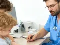 Lekarz bada małe dziecko, które trzyma mama. Na biurku leży dokumentacja medyczna, a lekarz notuje wyniki badania. CRP u noworodka jest ważnym wskaźnikiem.