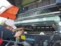 Rolnik w czapce z daszkiem pracuje przy prasie, która owija balot zieloną siatką. To praca przy maszynie rolniczej, być może marki rivierre casalis.