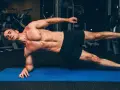Un homme musclé réalise un gainage planche sur un tapis bleu dans une salle de sport.