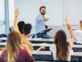 Uśmiechnięty wykładowca z książką w ręku wskazuje na studentów, którzy podnoszą ręce. Czy przed lecz stawiamy przecinek?