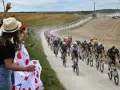 Spectators cheer as a peloton races on a dusty road during the Tour de France watch. Cyclists in colorful jerseys kick up dust in a dramatic scene.