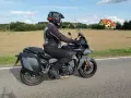 Motocyklista na szarym motocyklu Yamaha Tracer 900, z kuframi, w kasku, jedzie drogą. W tle pola i domy. Yamaha Tracer 900 dane techniczne.