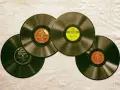 Vier alte Schallplatten liegen auf einer Leinwand. Eine davon hat ein gelbes MGM-Label, die anderen sind von Victrola und Victor. Sie erinnern an die Anfänge des Rocken Roll.