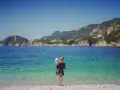 Kobieta z dzieckiem na rękach podziwia piękne Korfu plaże. Woda jest krystalicznie czysta, a w oddali widać góry.