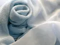 Miękka, błękitna bawełna merceryzowana zwinięta w kształt róży, obok delikatnego, białego woalu.
