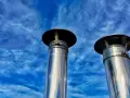 Due camini in acciaio inox, modello uni 7129 scarico a parete, brillano sotto un cielo blu con nuvole sottili.