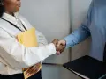 Uścisk dłoni po rozmowie o przedłużenie umowy o pracę. Kobieta trzyma żółtą teczkę, mężczyzna czarną.
