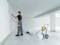 Man i skyddsutrustning sprutmålar en vägg med en färgspruta. En maskin och en hink står på plastskydd.