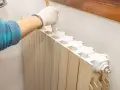 Mano con guanto bianco che dipinge un termosifone beige con vernice bianca, pronto per imbiancare la stanza.