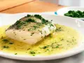 Un filete de merluza en salsa verde, con perejil picado, servido en un plato blanco.