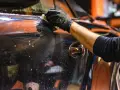 Mano con guanto nero pulisce la carrozzeria lucida di un'auto, rimuovendo la resina con un panno umido e schiumoso.