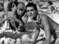Dwóch młodych mężczyzn na plaży, jeden w czapce, drugi z chustą na szyi.