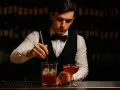 Barman w eleganckim stroju miesza drinka z lodem w szklanicy, obok stoi gotowy koktajl.