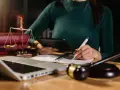 Prawniczka analizuje dane na tablecie, notując w zeszycie. Obok leżą waga sprawiedliwości, młotek sędziowski i laptop. Pomaga to zrozumieć, jak sprawdzić przedawnienie długu.