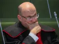 Sędzia w todze z czerwoną lamówką, w okularach, z ręką na brodzie, zamyślony.