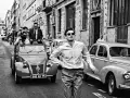 Un uomo con sigaretta in bocca, occhiali da sole e camicia a righe, in strada con auto d'epoca, evoca lo spirito de **la nouvelle vague**.