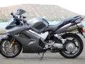 Szary motocykl Honda VFR 800 z widokiem na wodę i wzgórze w tle.