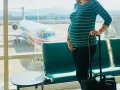 Ciężarna kobieta z walizką czeka na lotnisku. W tle widać samolot i pasażerskie fotele. Latanie samolotem w ciąży to wyzwanie, ale podróż dobiega końca.