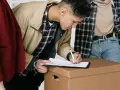 Joven firma contratos de alquiler sobre una caja de mudanza, acompañado de dos personas.
