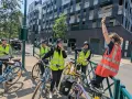 Des adultes souriants, casqués et en gilets fluo, prêts à apprendre à faire du vélo adulte. Ils sont avec leurs vélos, devant un immeuble moderne.