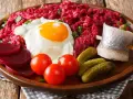 Herzhaftes Labskaus essen: Rote Bete, Spiegelei, Matjes, Cornichons und Tomaten auf einem Holzteller.