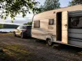 En husvagn och bil parkerad vid en sjö, redo för en husvagnssemester i Sverige. Solen lyser på den öppna dörren.