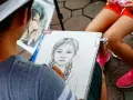 Artysta tworzy portret dziewczynki z warkoczami. Obok leży gotowy rysunek chłopca w okularach.