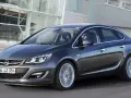 Ciemnoszary Opel Astra J 1.6 benzyna w ruchu, z kierowcą w okularach za kierownicą, na tle nowoczesnego budynku.