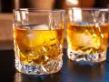 Zwei Gläser mit bernsteinfarbenem on the rocks drink, beleuchtet von warmem Licht.