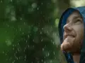 Mann mit Bart blickt lächelnd in den Regen, seine blaue Regenjacke hält ihn trocken.