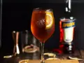 Orzeźwiający Aperol Spritz z plasterkiem pomarańczy, obok butelka Aperol. Idealny wybór zamiast Hugo.
