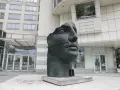 Okaleczona, ciemna rzeźba głowy z opaską na ustach, na tle nowoczesnego budynku.
