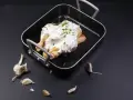 Un poulet entier est nappé de fromage blanc crémeux et garni d'herbes fraîches dans un plat noir. Des gousses d'ail sont disposées autour.