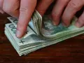 Dłonie przewracają stos banknotów 100 zł na drewnianym blacie.