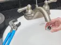 Hand holding a removed faucet aerator with red buildup, next to pliers and duct tape on a sink.