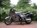 Srebrny motocykl Kawasaki GTR 1000 zaparkowany na czerwonej kostce brukowej, otoczony zielenią.