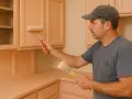 Uomo con cappellino dipinge mobili da cucina in legno con pittura murale color pesca, usando rullo e pennello.