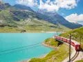 A red train winds along a track beside a vibrant turquoise lake, with lush green mountains and a bright blue sky in the background. This is a classic como to switzerland train journey.